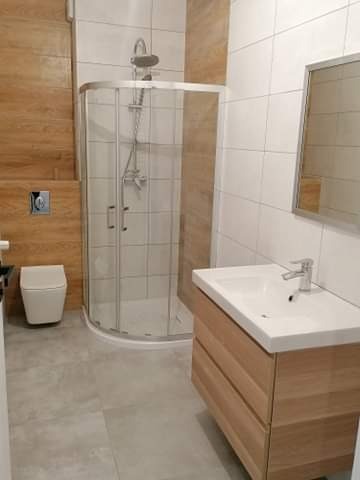 Nowoczesna łazienka z kabiną prysznicową z zaokrąglonym szkłem, umywalką z szafką z jasnego drewna i płytkami w odcieniach bieli i brązu.