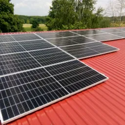 Panele słoneczne na czerwonym blaszanym dachu budynku z widokiem na zielone drzewa i pole w tle.