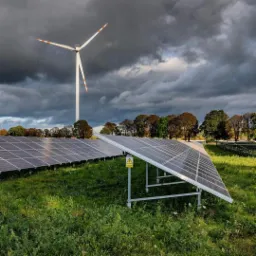 Rząd paneli słonecznych w polu z wysoką trawą, w tle turbina wiatrowa i ciemne chmury burzowe. Żółty znak ostrzegawczy na słupku przy panelach.