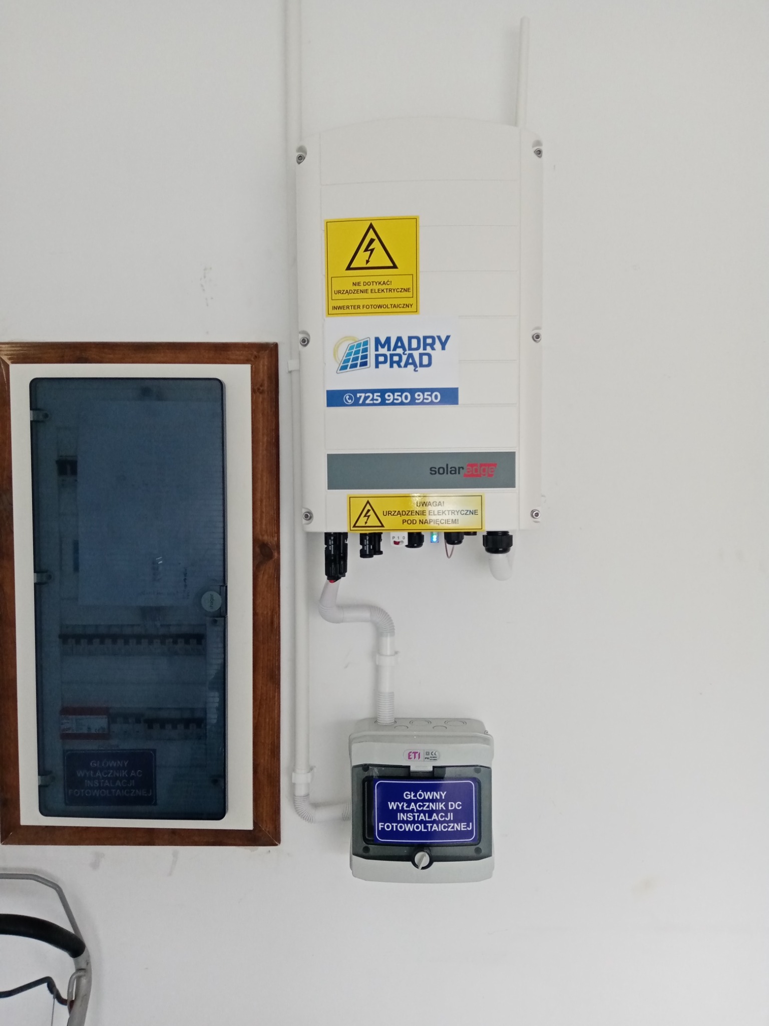 Inwerter fotowoltaiczny SolarEdge zamontowany na ścianie obok skrzynki z wyłącznikiem AC i wyłącznikiem DC, widoczne ostrzeżenia o napięciu.