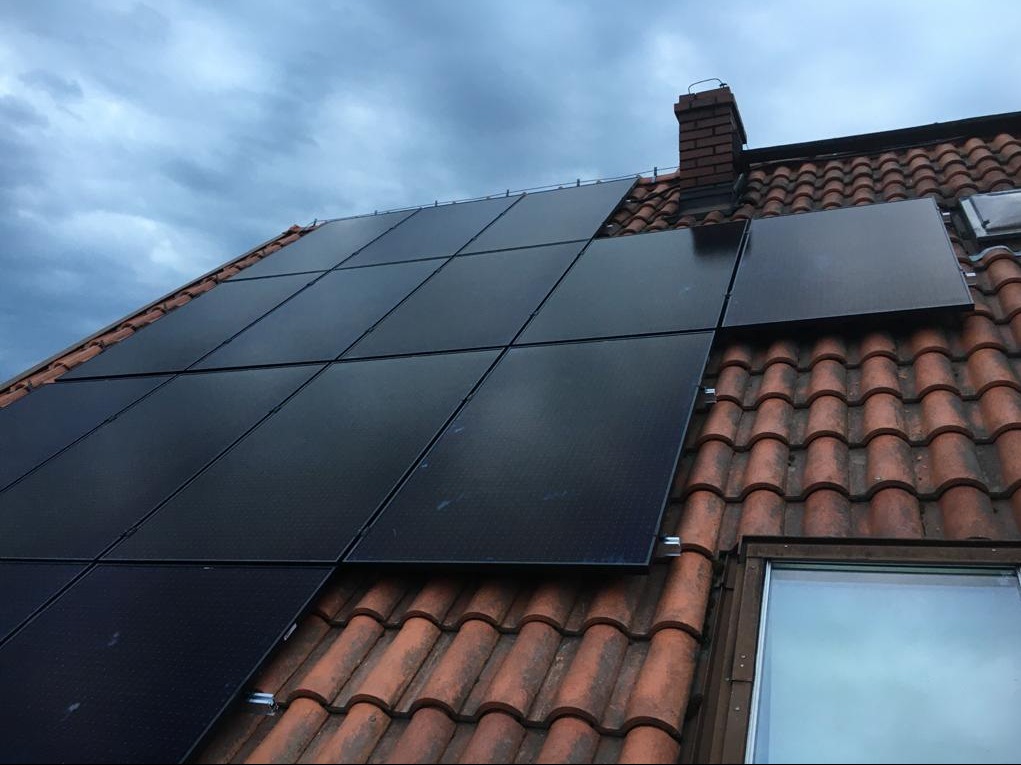 Panele fotowoltaiczne zamontowane na dachu pokrytym czerwoną dachówką, widoczny komin i okno dachowe, pochmurne niebo w tle.