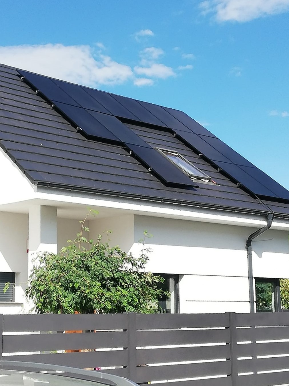 Czarny dach z panelami słonecznymi na nowoczesnym, białym domu jednorodzinnym z oknem dachowym, widoczne zza grafitowego ogrodzenia i zieleni.