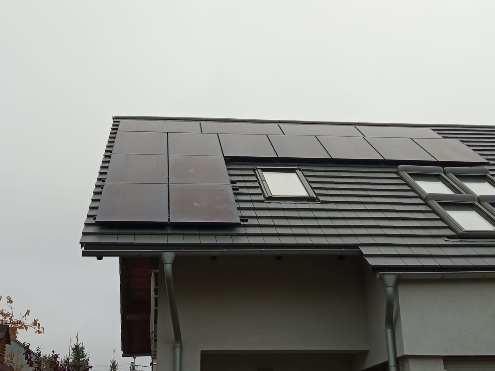 Częściowo zamontowane panele słoneczne na dachu domu z dachówką ceramiczną i oknami dachowymi, widok z dołu w pochmurny dzień.