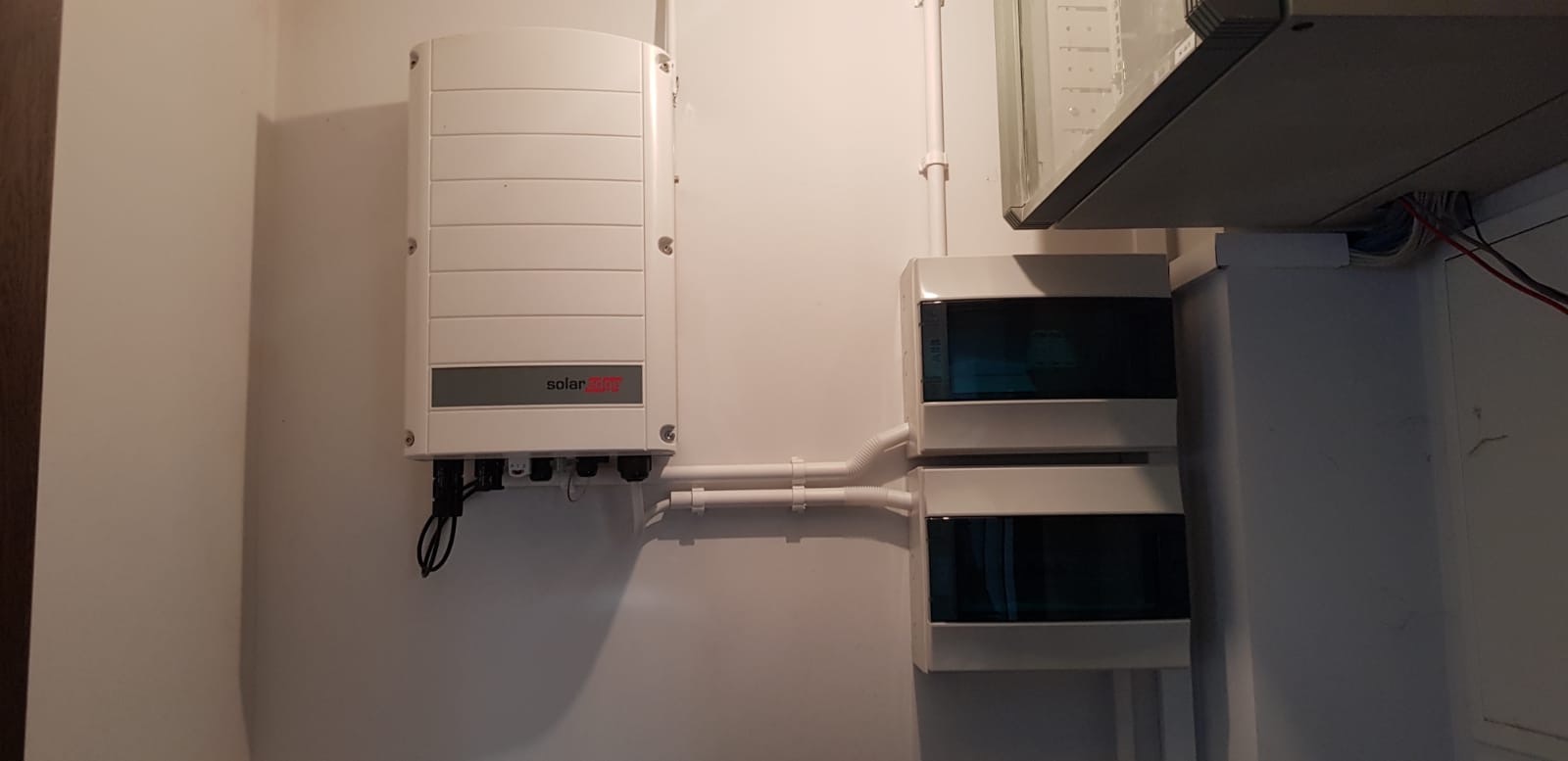 Biały falownik SolarEdge zamontowany na ścianie obok dwóch skrzynek bezpiecznikowych z niebieskimi szybkami, połączonych białymi rurkami PCV.
