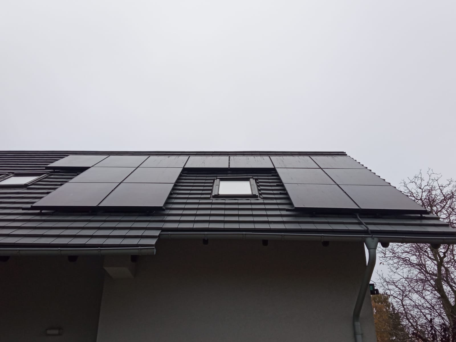 Czarne panele słoneczne zamontowane na ciemnoszarym dachu domu jednorodzinnego z oknem dachowym, widok z dołu w pochmurny dzień.