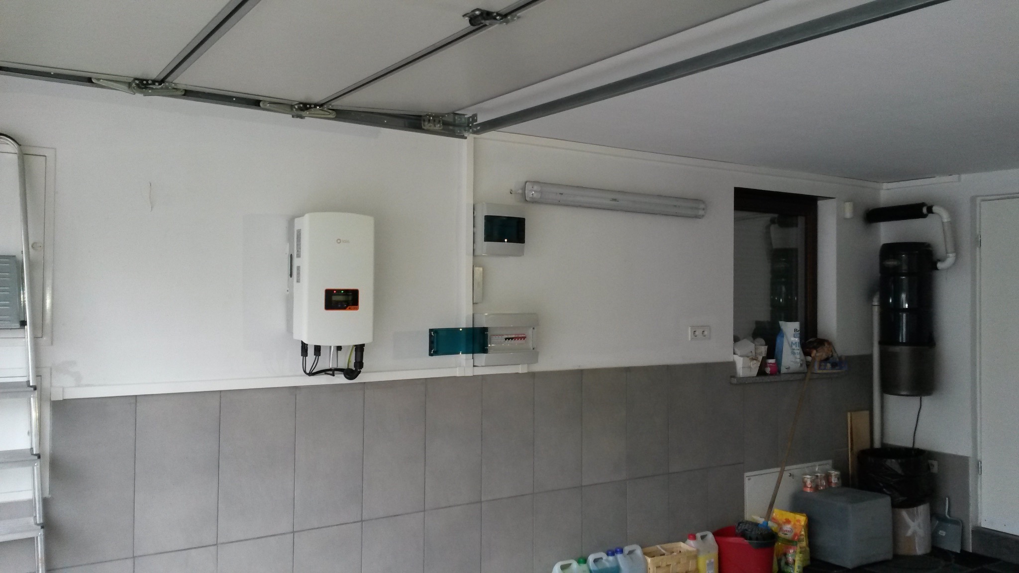 Biały falownik fotowoltaiczny zamontowany na ścianie garażu z szarą glazurą, obok skrzynka elektryczna i okno.