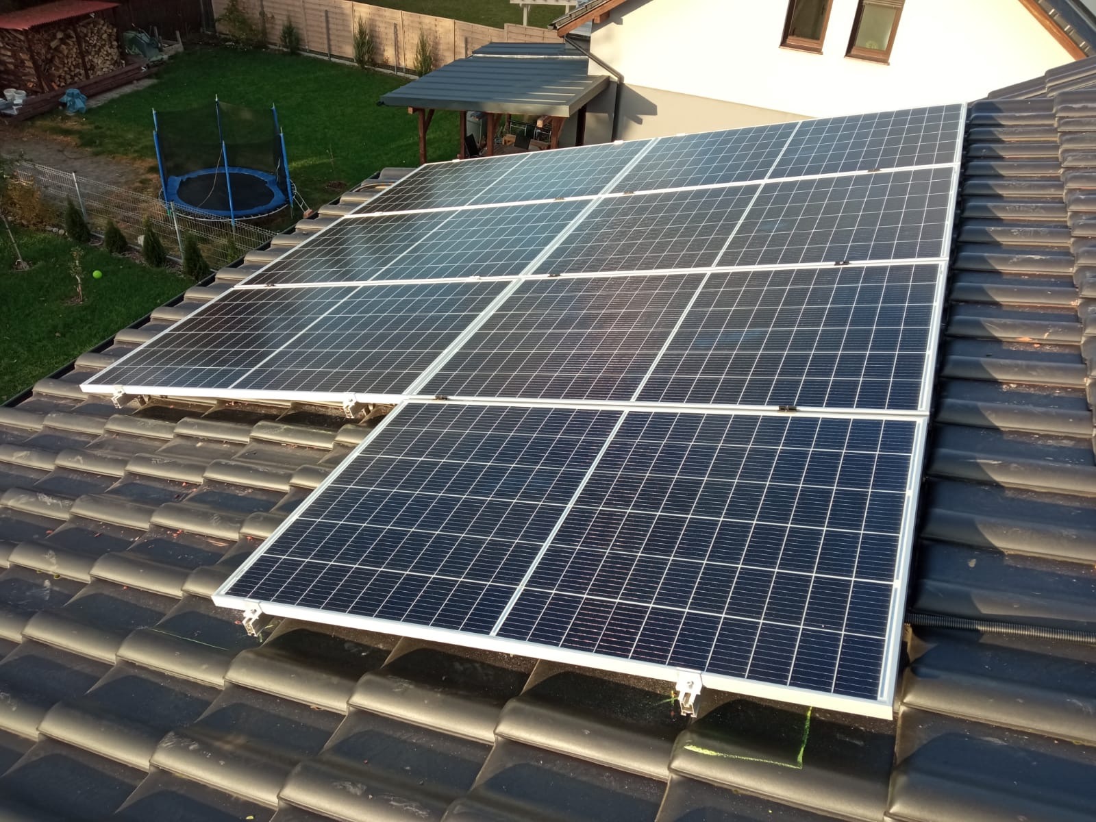 Panele słoneczne zamontowane na ciemnym, ceramicznym dachu domu jednorodzinnego, widok z góry, w tle trawnik z trampoliną.