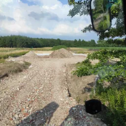 Widok z kabiny maszyny budowlanej na plac z gruzem i wykopami, w tle drzewa i błękitne niebo z chmurami.
