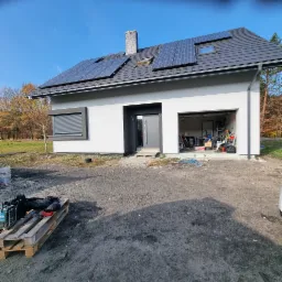 Nowoczesny, parterowy dom z szarą stolarką okienną i drzwiową oraz panelami słonecznymi na dachu, w trakcie prac wykończeniowych na zewnątrz, z widocznym bałaganem na podwórku i paletą z narzędziami.