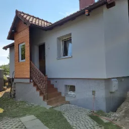 Dom jednorodzinny po remoncie elewacji, z drewnianą dobudówką nad wejściem i nowymi schodami, ściany w odcieniach szarości, widoczny fragment chodnika z kostki brukowej i trawnika.