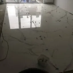 Układanie jasnych, marmurkowych płytek podłogowych z użyciem krzyżyków dystansowych, widoczny pojemnik z zaprawą klejową.