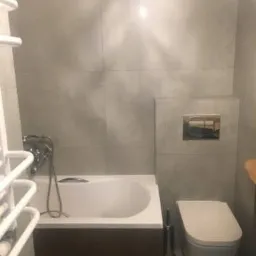 Nowoczesna łazienka z wanną obudowaną ciemnym drewnem, wiszącą toaletą i grzejnikiem drabinkowym, wykończona szarymi płytkami imitującymi beton.