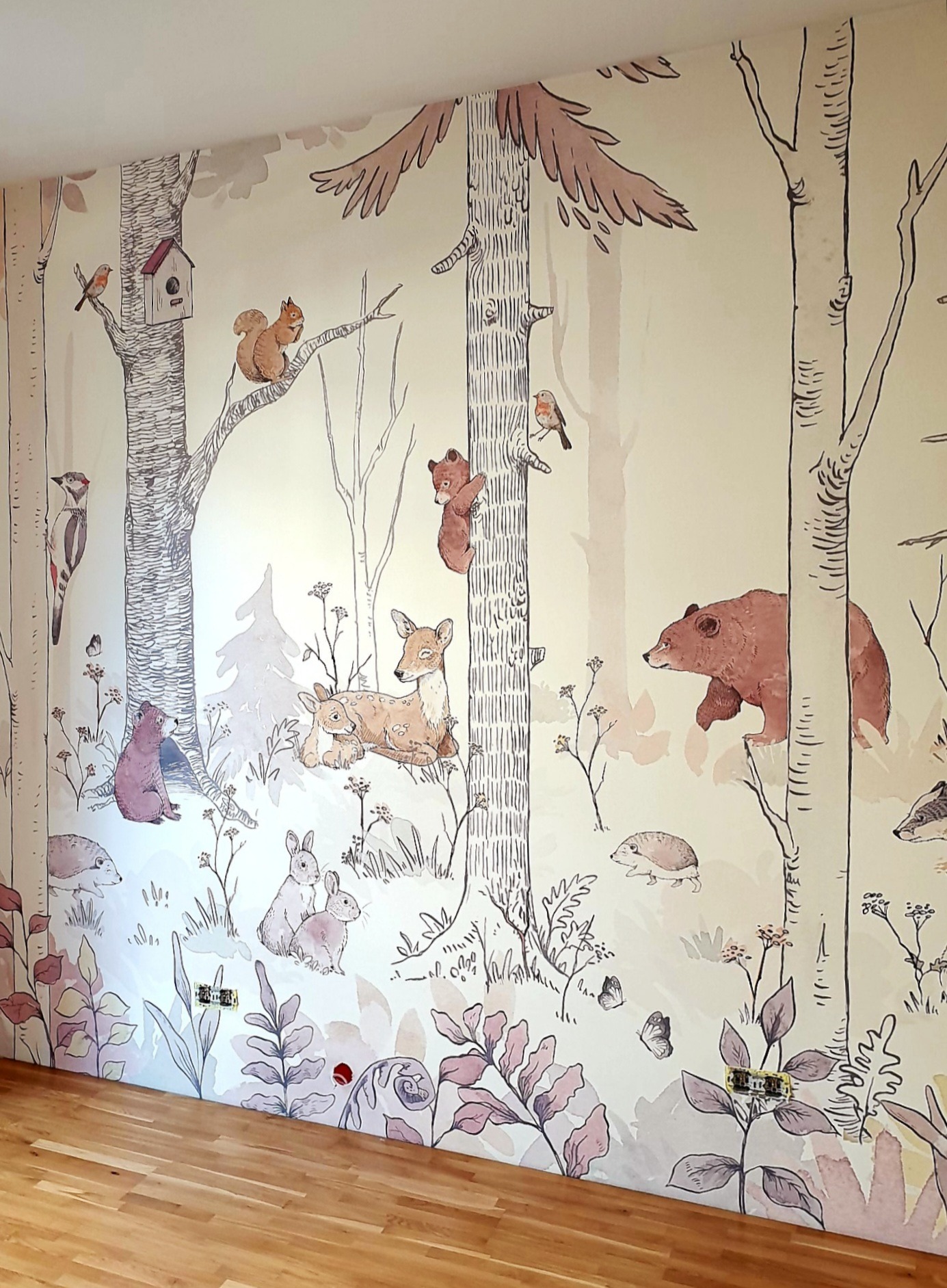 Ściana w pokoju dziecięcym z ręcznie malowaną tapetą przedstawiającą leśny krajobraz z motywami zwierząt: niedźwiedzie, sarny, wiewiórki, ptaki i jeże, na tle drzew i roślin w pastelowych kolorach.