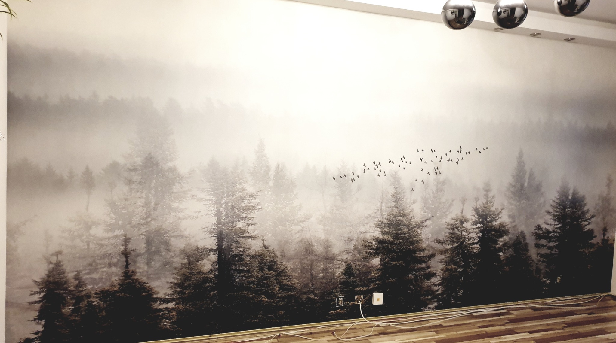Fototapeta z motywem mglistego lasu iglastego i przelatujących ptaków, zamontowana na ścianie w jasnym pokoju z drewnianą podłogą.