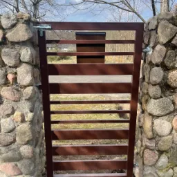 Brązowa metalowa brama z poziomymi szczeblami, zamontowana między kamiennymi słupkami, z widoczną zasuwką i skrzynką na listy, na tle trawnika i drzew bez liści.