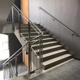 Schody wykończone szarą płytką z pionowymi, metalowymi barierkami i pojedynczą poręczą na ścianie; na górze drzwi ewakuacyjne z zielonym znakiem.