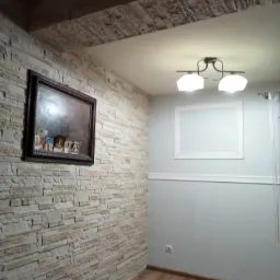 Pokój po odświeżeniu: ściana z dekoracyjnej okładziny imitującej kamień, drewniana podłoga, szara ściana z białym, ozdobnym obramowaniem i ramą, lampa sufitowa z dwoma kloszami.