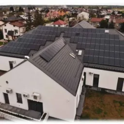 Widok z góry na biały budynek z zamontowanymi panelami fotowoltaicznymi na dachu, z widocznymi klimatyzatorami na ścianach i zabudową osiedlową w tle.