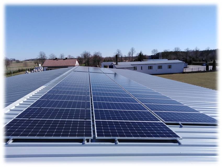 Rząd paneli słonecznych na dachu z blachy trapezowej, widok z góry, w tle budynki gospodarcze i drzewa.