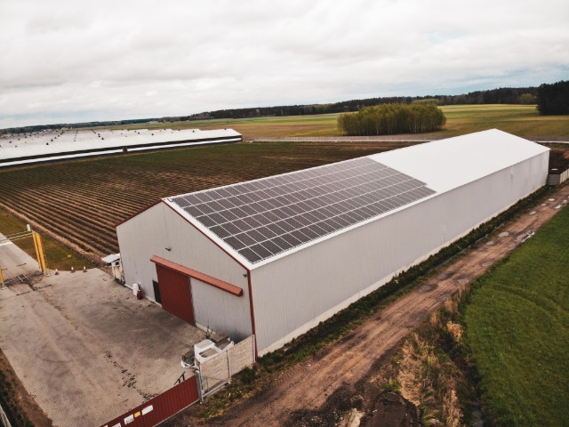 Widok z drona na duży, szary budynek gospodarczy z częściowo zamontowanymi panelami słonecznymi na dachu, otoczony polami i lasem w oddali.