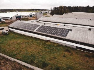 Widok z lotu ptaka na budynki gospodarcze z panelami słonecznymi na dachu, otoczone zielenią i lasem w tle.