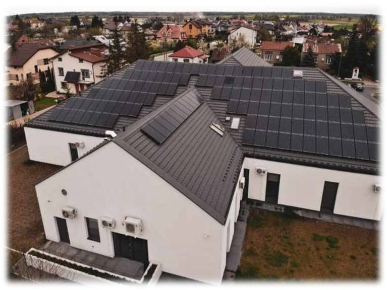 Widok z góry na biały budynek z zamontowanymi panelami fotowoltaicznymi na dachu, z widocznymi klimatyzatorami na ścianach i zabudową osiedlową w tle.