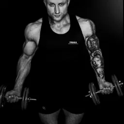 Czarno-białe zdjęcie muskularnego mężczyzny z tatuażami, w czarnym podkoszulku z logo, trzymającego hantle na czarnym tle.