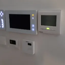 Panel sterowania inteligentnego domu z wyświetlaczami temperatury, interkomem i włącznikami światła, zamontowany na białej ścianie.