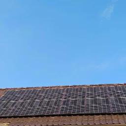 Panele fotowoltaiczne zamontowane na brązowym, spadzistym dachu pokrytym dachówką, widoczne pod błękitnym niebem. Część dachówek usunięta podczas montażu paneli.