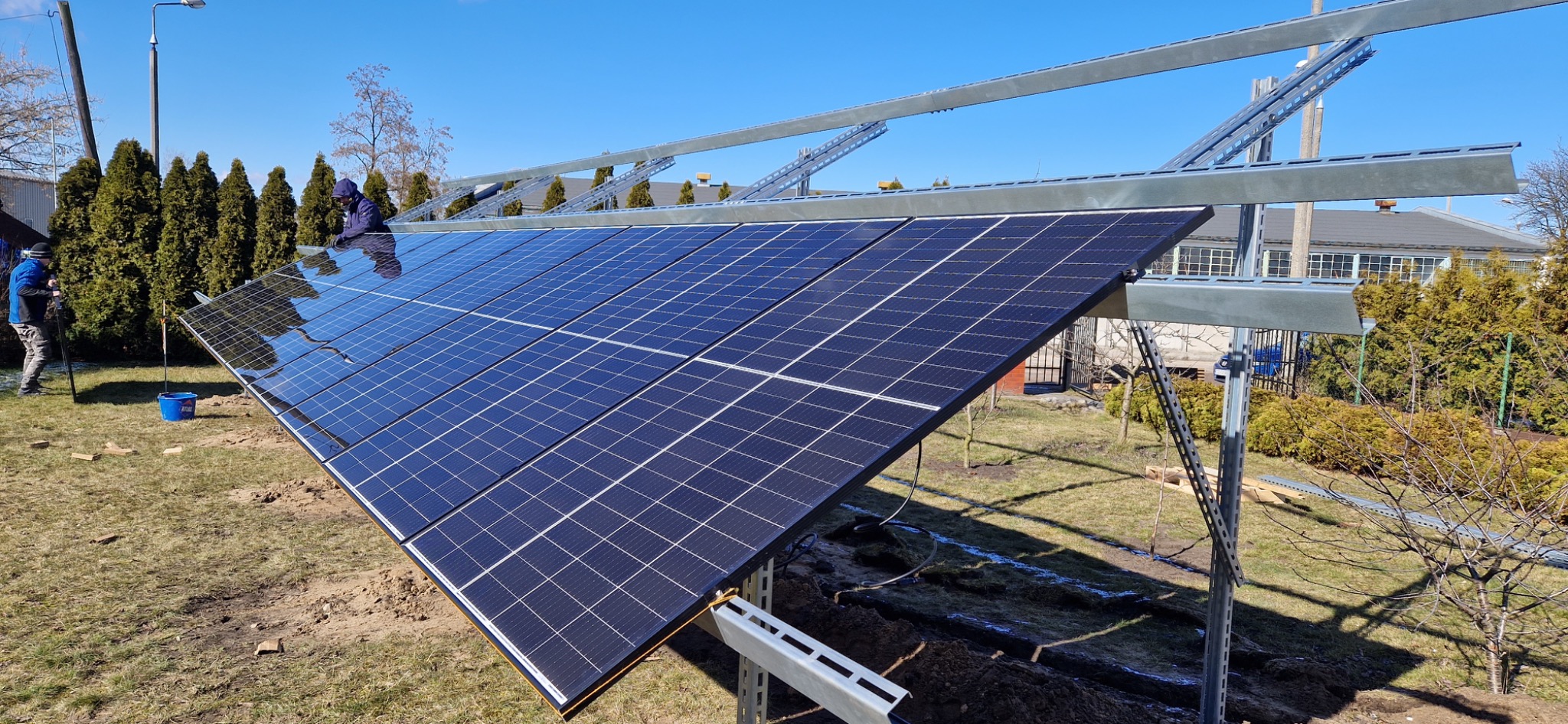 Montaż paneli fotowoltaicznych na metalowej konstrukcji w ogrodzie, widoczni pracownicy w odzieży roboczej, błękitne niebo.