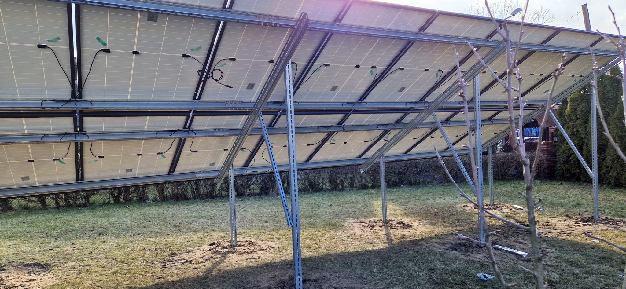 Widok od spodu na panele fotowoltaiczne zamontowane na metalowej konstrukcji na trawniku, widoczne kable i elementy mocujące, w tle krzewy i drzewa.