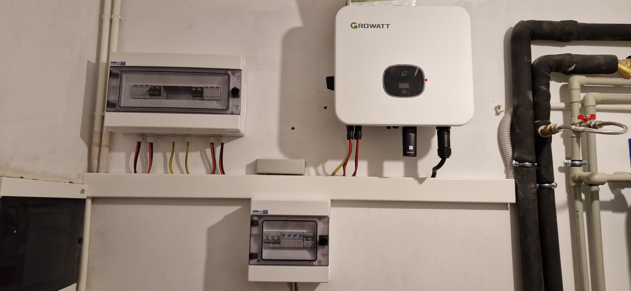 Biały inwerter Growatt zamontowany na ścianie obok dwóch skrzynek elektrycznych z widocznymi przewodami i rurami instalacji grzewczej.