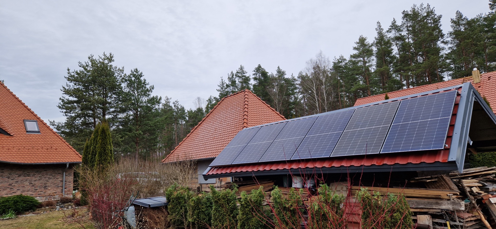 Panele fotowoltaiczne zamontowane na dachu budynku z czerwoną dachówką, widok z poziomu gruntu, w tle drzewa iglaste i budynki.