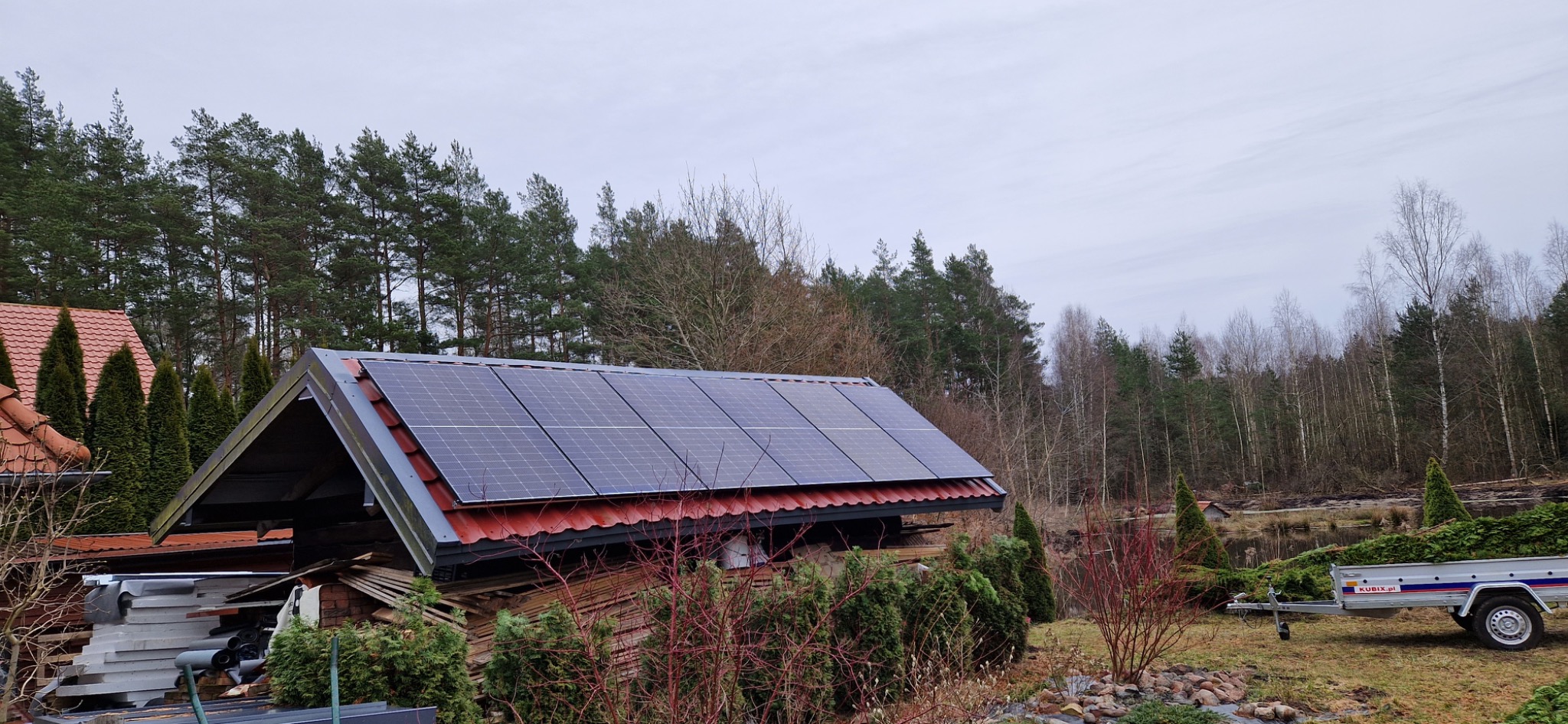Panele fotowoltaiczne na dachu budynku gospodarczego z czerwonej dachówki, w tle las i przyczepa z zielonymi tujami.