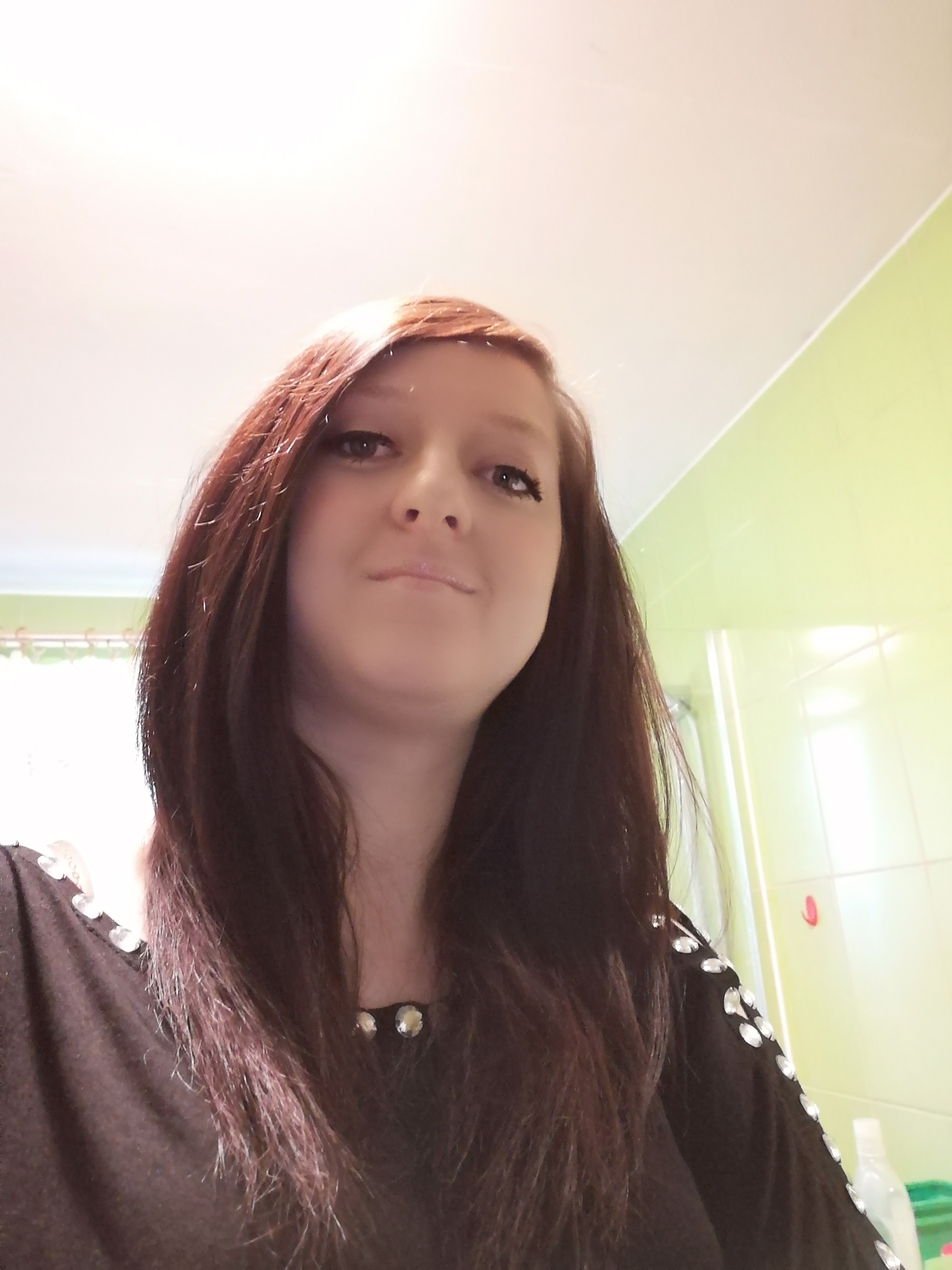 Portret kobiety z długimi, ciemnymi włosami w łazience z zielonymi płytkami, ubrana w czarną bluzkę z aplikacjami z kryształków.