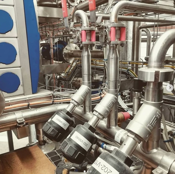 Złożona instalacja rur ze stali nierdzewnej z zaworami i czujnikami w zakładzie przemysłowym, widoczne oznaczenia na elementach.