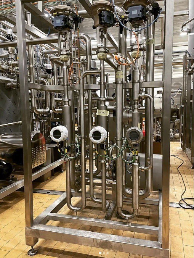 Skomplikowana instalacja rurowa ze stali nierdzewnej z zaworami automatycznymi i czujnikami, umieszczona na ramie na tle wykafelkowanej podłogi przemysłowej.