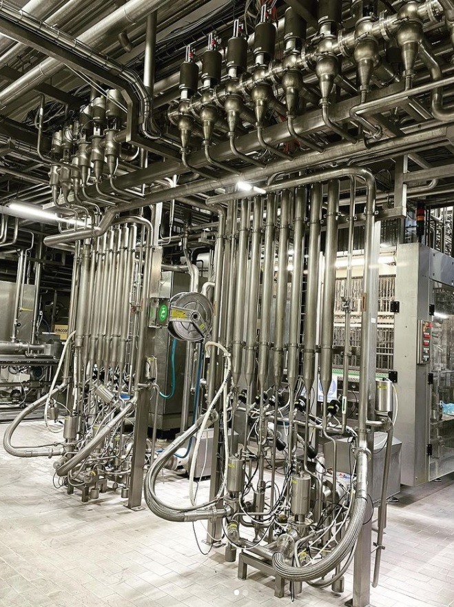 Kompleksowy system rur ze stali nierdzewnej w zakładzie przemysłowym, widoczne liczne zawory i połączenia, oświetlenie górne.