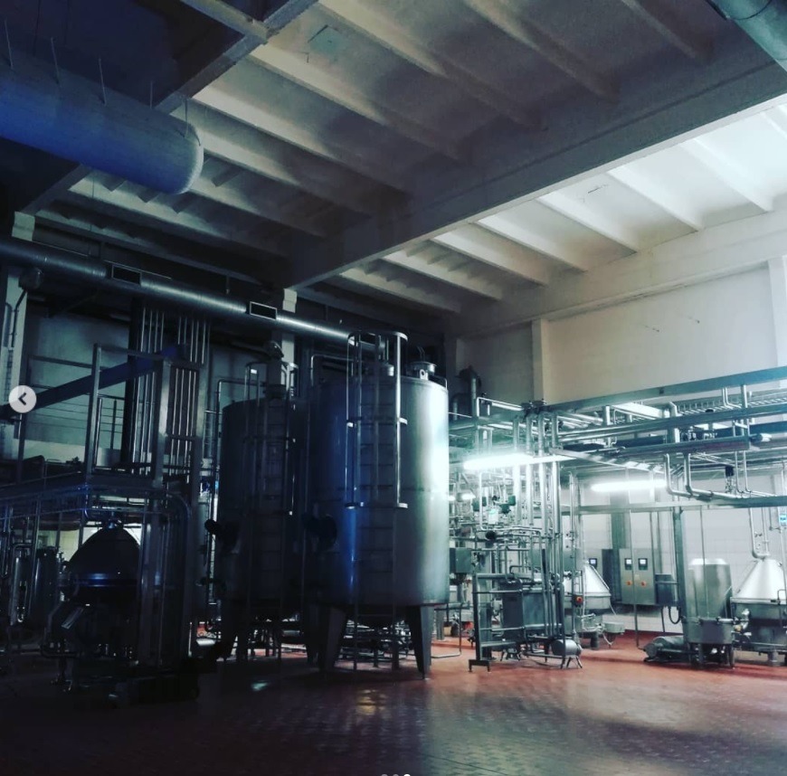 Wnętrze hali produkcyjnej z instalacją rurociągów i zbiorników ze stali nierdzewnej na czerwonej posadzce, oświetlone światłem jarzeniowym.