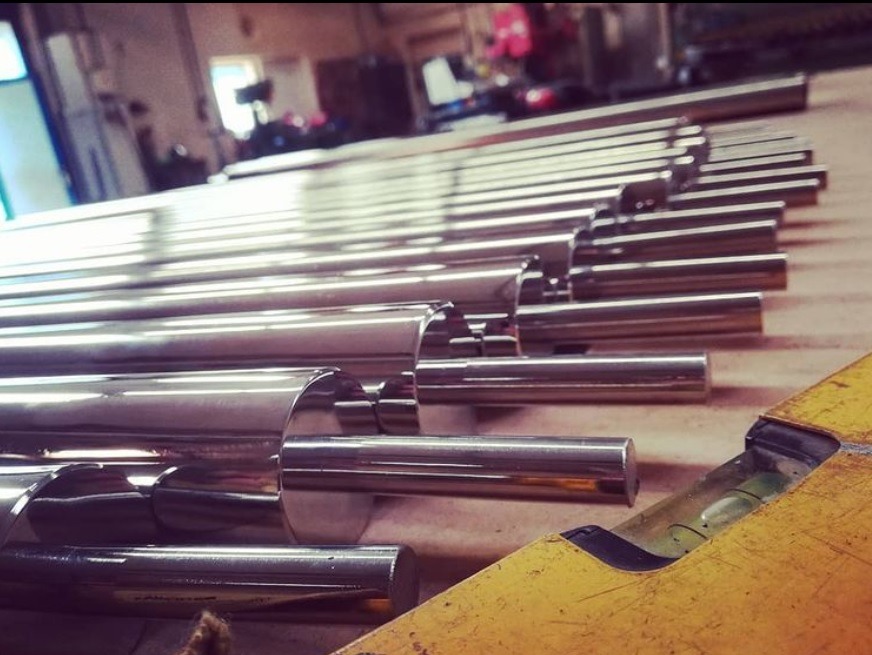 Polerowane rury ze stali nierdzewnej ułożone równolegle na żółtym stole warsztatowym, w tle widoczne elementy hali.