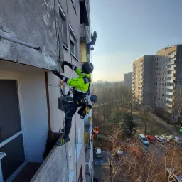 Pracownik w uprzęży, w kasku i jaskrawej kurtce, zawieszony na linie, skrobie elewację bloku mieszkalnego, w tle widoczne inne budynki i drzewa.