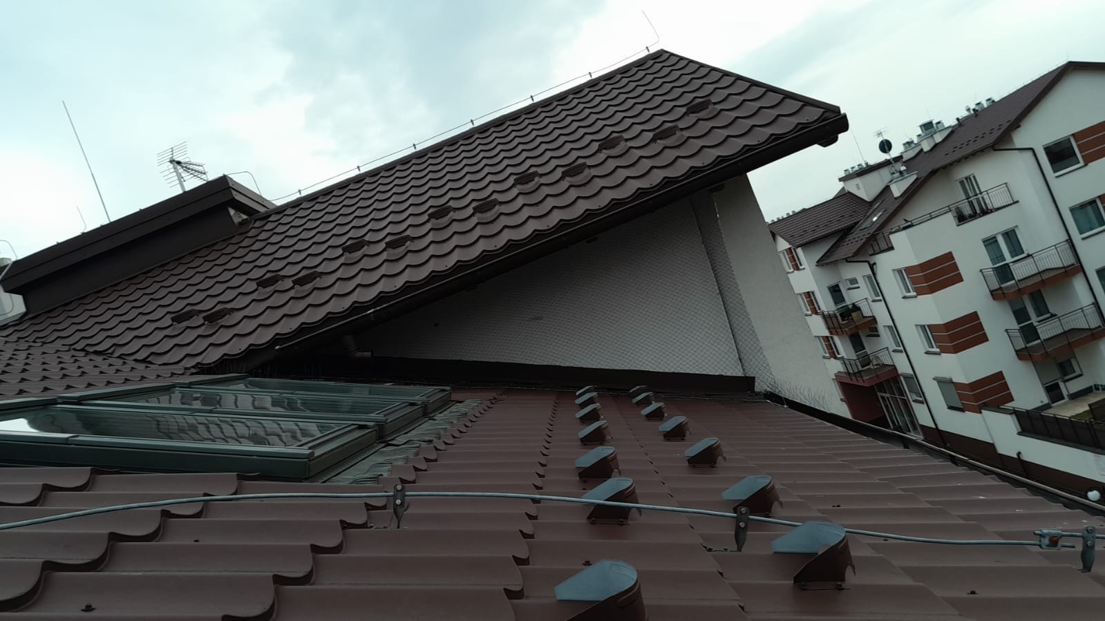 Brązowy dach z dachówki ceramicznej z oknami dachowymi i systemem odgromowym, widok z góry, w tle budynek mieszkalny.