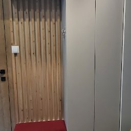 MPL Meble na wymiar - Szara szafa wnękowa z drewnianym panelem i siedziskiem z bordową poduszką, dopasowana do wnętrza, widoczna na tle podłogi z płytek imitujących drewno.