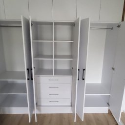 MPL Meble na wymiar - Biała szafa wnękowa z otwartymi drzwiami, widoczne półki, szuflady i drążki na ubrania. Minimalistyczny design, jasne wnętrze, drewnopodobna podłoga.