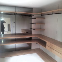 MPL Meble na wymiar - Kremowo-brązowa garderoba z lustrami i półkami, widoczna w jasnym pomieszczeniu. W lustrze odbija się sylwetka osoby robiącej zdjęcie.