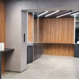 MPL Meble na wymiar - Nowoczesna łazienka z drewnianymi szafkami, białymi umywalkami i grafitowymi blatami. Lustra z podświetleniem LED. W tle widoczny napis na ścianie.