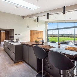 MPL Meble na wymiar - Nowoczesna kuchnia z wyspą i drewnianym blatem, minimalistyczne krzesła barowe, duże okno z widokiem na zieleń. Oświetlenie punktowe i podłużne lampy wiszące.