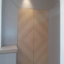 Dwudrzwiowa szafa wnękowa z drewnianymi frontami ułożonymi w jodełkę, otoczona szarymi panelami. Górne podświetlenie podkreśla teksturę drewna i geometryczny wzór.