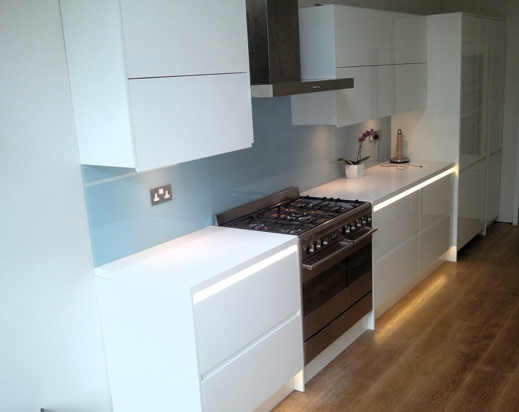 Nowoczesna, biała kuchnia z podświetleniem LED pod blatem, stalowym okapem i płytą gazową. Jasne drewno na podłodze. Minimalistyczny design.