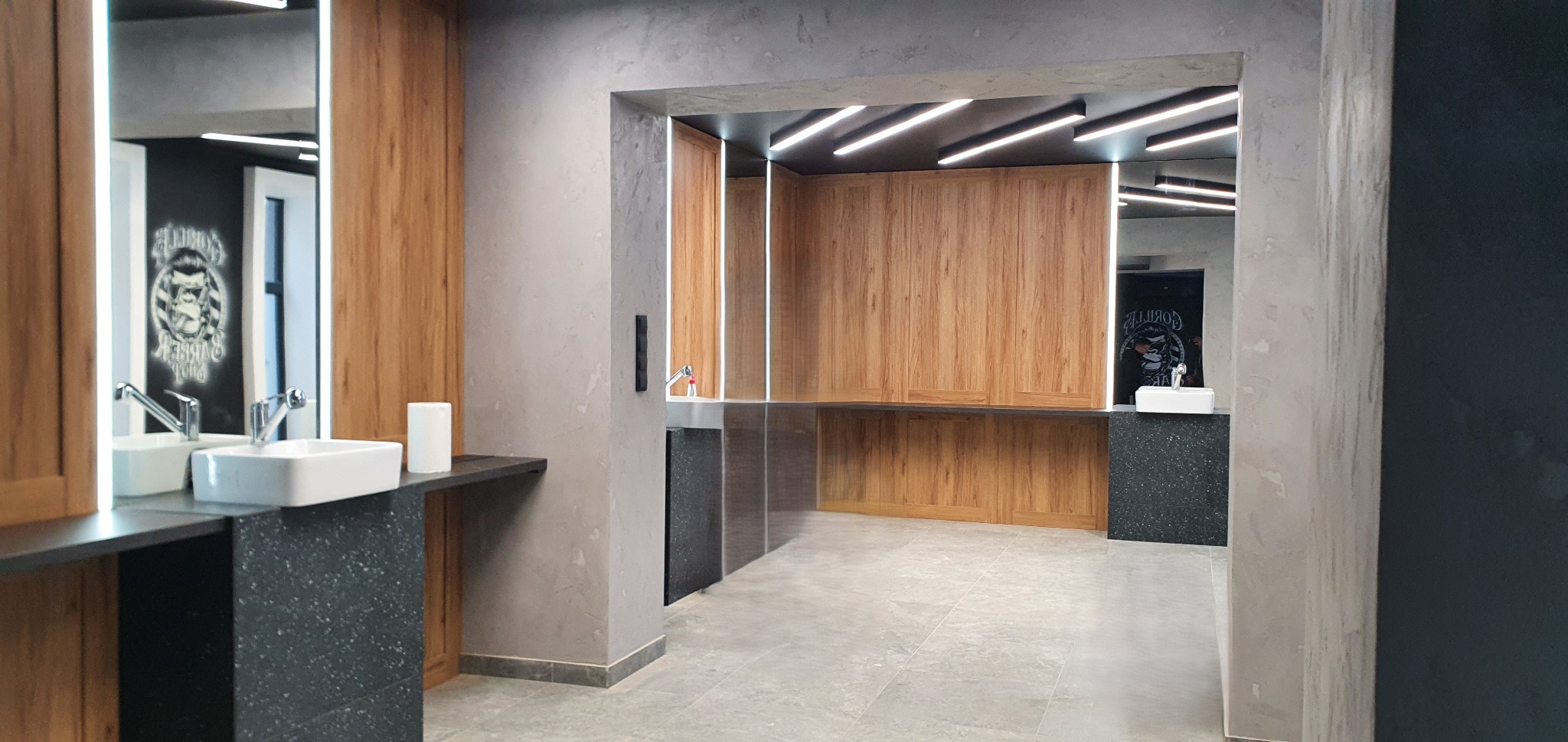 Nowoczesna łazienka z drewnianymi szafkami, białymi umywalkami i grafitowymi blatami. Lustra z podświetleniem LED. W tle widoczny napis na ścianie.